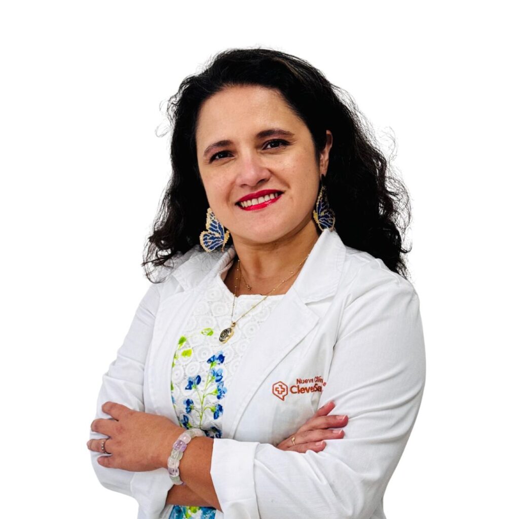 Portrait of a female healthcare professional in a white coat, smiling with arms crossed, wearing blue patterned earrings and a floral blouse visible underneath.