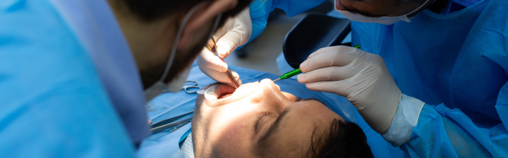 Dentists in blue scrubs perform a dental procedure on a patient, using surgical tools over the open mouth