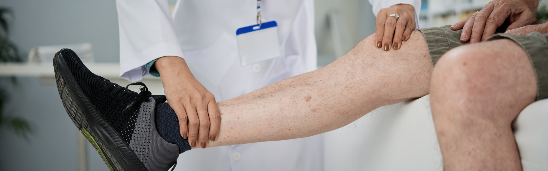 Medical professional examines a patient's leg, supporting the shin during a check-up with a blue ID badge visible on white coat.
