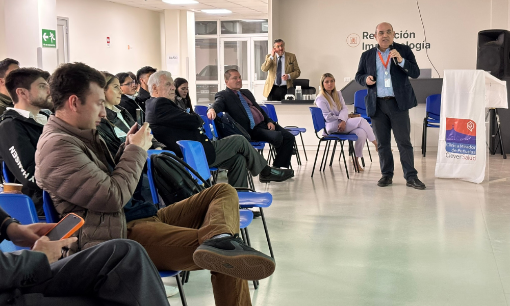 Professional speaker in a blazer addresses an audience in a conference room, with attendees seated on blue chairs and a podium to the side.
