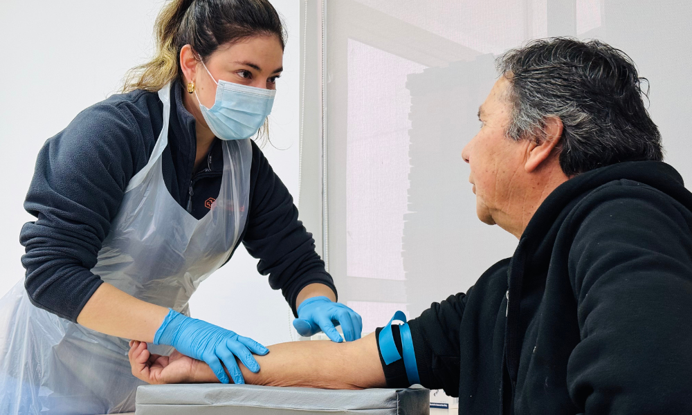 Female nurse wearing a mask, gloves, and a plastic apron, drawing blood from a man's forearm with a tourniquet visible.