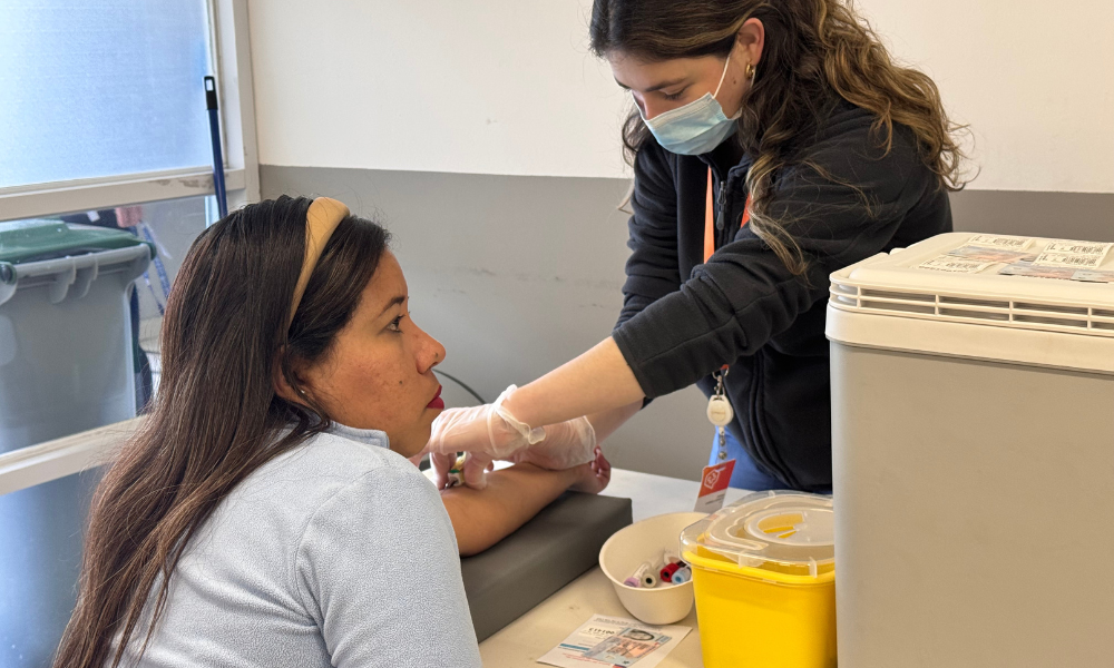 Nurse wearing a mask and gloves draws blood from a seated woman's arm in a clinic.