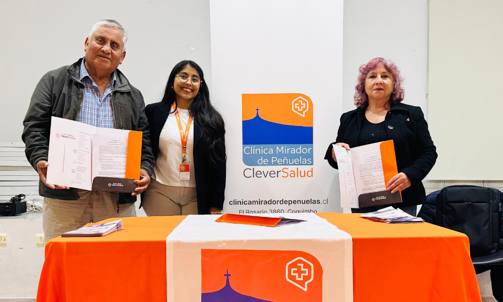 Three people stand behind an orange-clad info table at a health clinic booth, each holding documents in front of a banner that reads Clinica Mirador de Penuelas/Clever Salud.