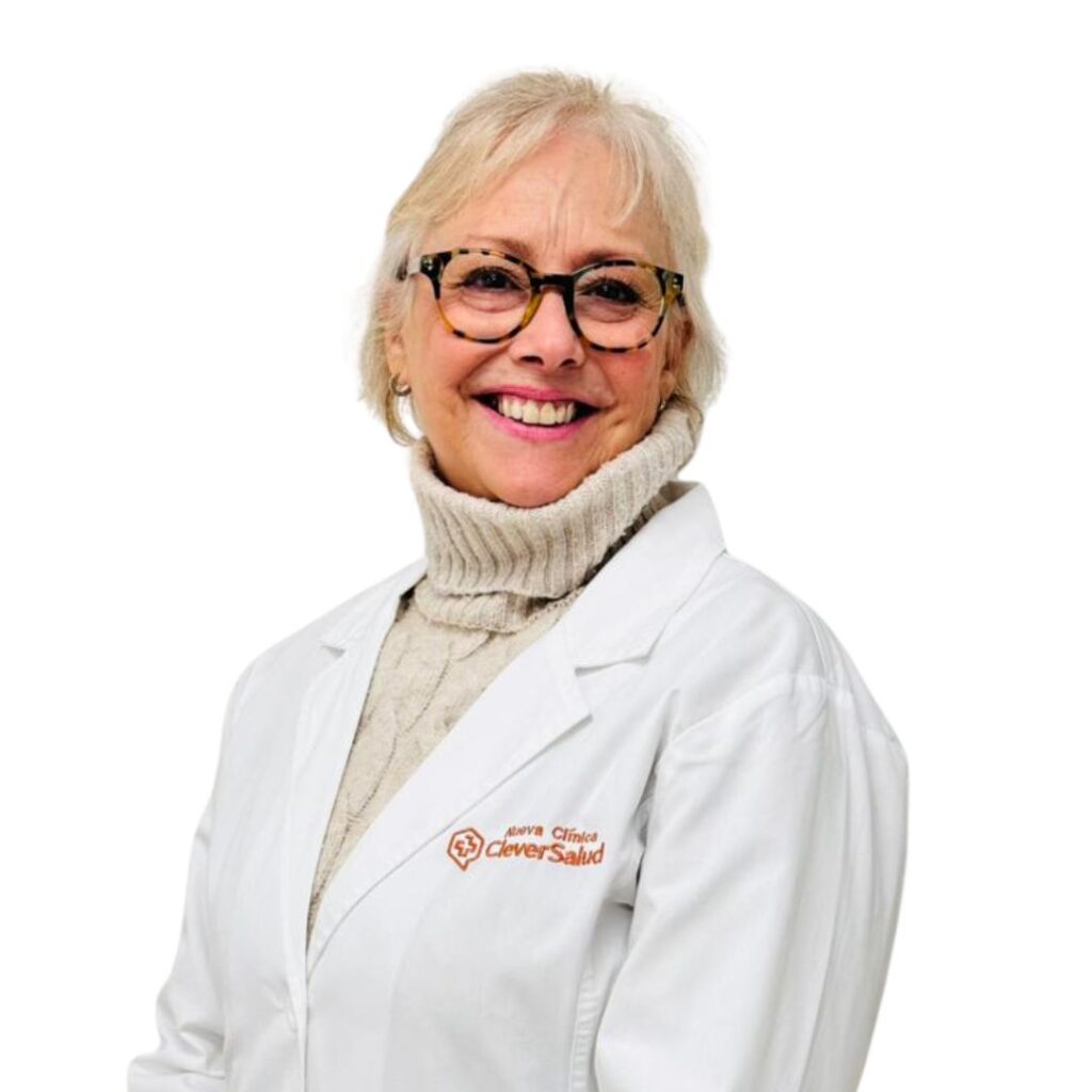 Smiling senior woman in a white lab coat and beige turtleneck, wearing tortoiseshell glasses, against a white background. The coat has an orange logo on the chest area.