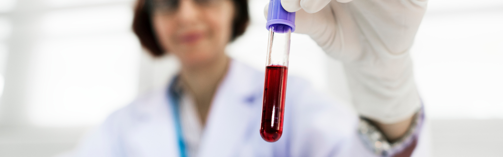 Gloved lab technician holds a blood-filled test tube with a purple cap toward the camera.