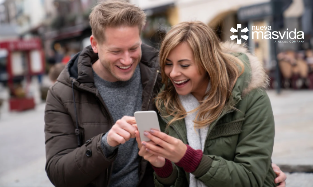 Man and woman in warm jackets smiling as they look at a smartphone on a city street; branding logo in the top-right corner.
