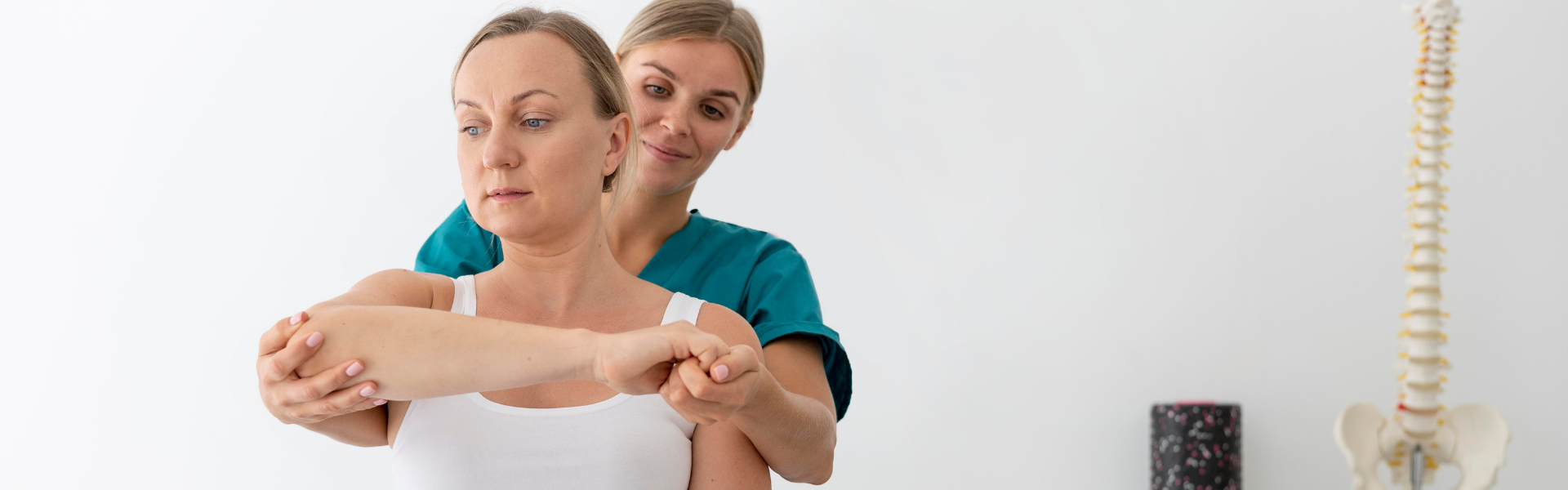 Therapist guides a patient through a shoulder stretch during physical therapy in a clinic, with a spine model visible in the background.