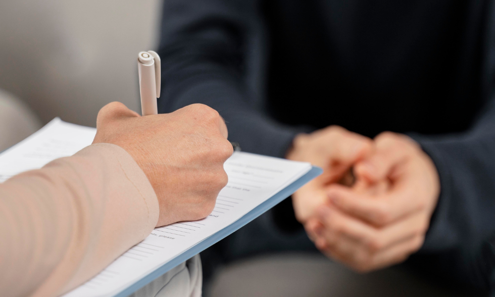 Person signs a document with a pen while the other person's hands are clasped in the background, suggesting a formal sign‑in or agreement moment.