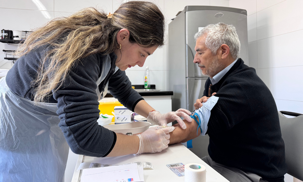 Healthcare worker draws blood from a man's arm in a clinical room, using gloves and a tourniquet.