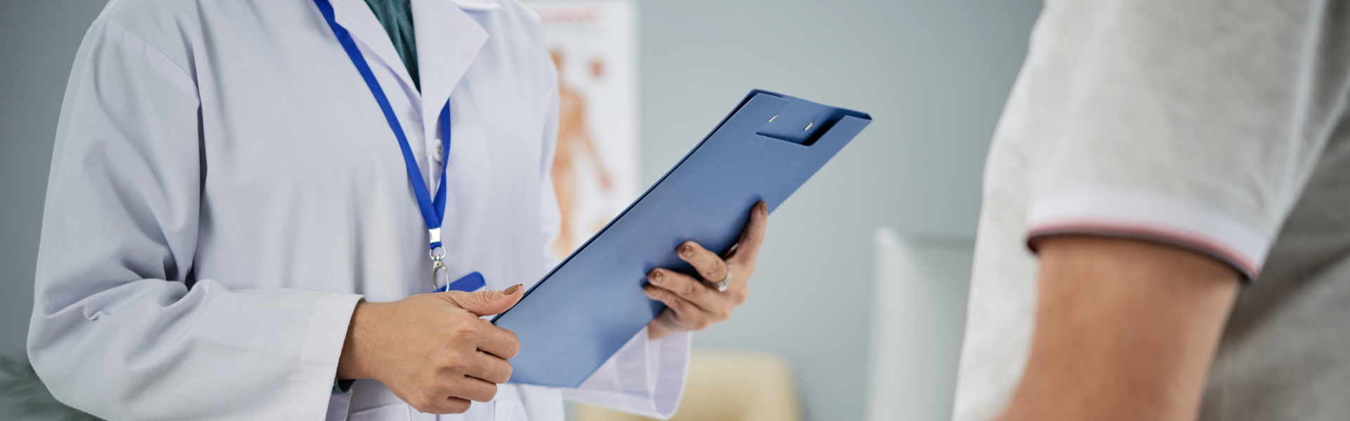 Healthcare professional in a white coat holds a blue clipboard and speaks with a patient in a clinic.