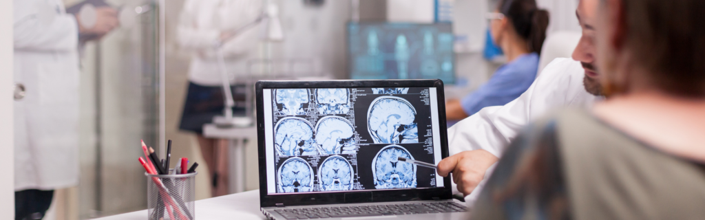 Medical professionals review brain MRI scans on a laptop, one pointing at the screen with a pointer.