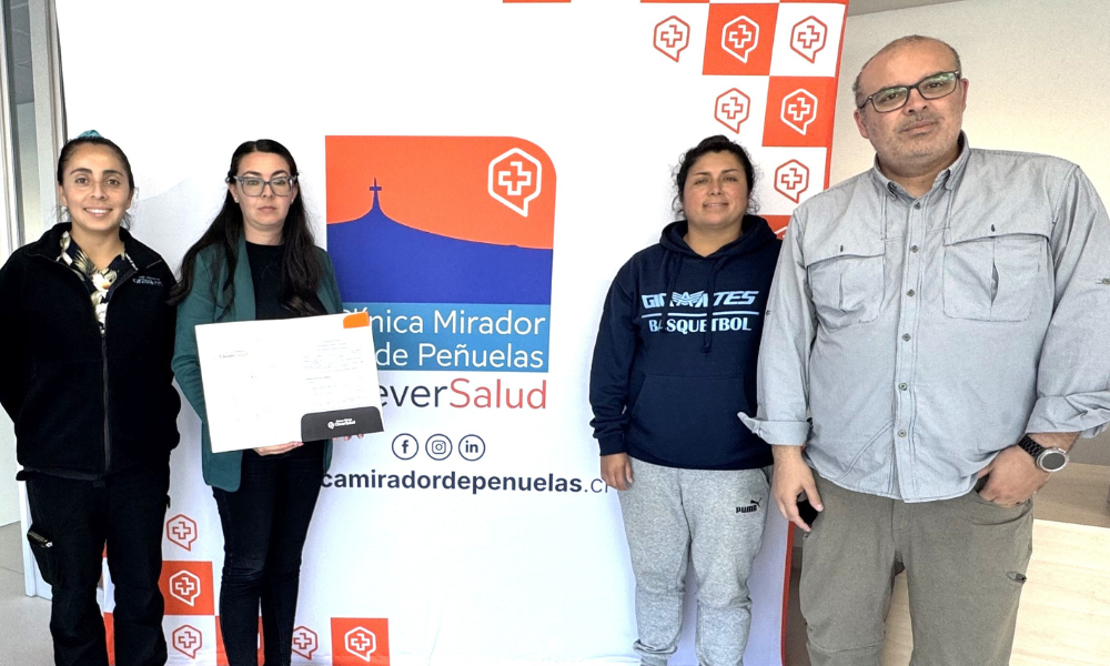 Four adults pose indoors in front of a white/orange banner with logos; the second person from the left holds a certificate.