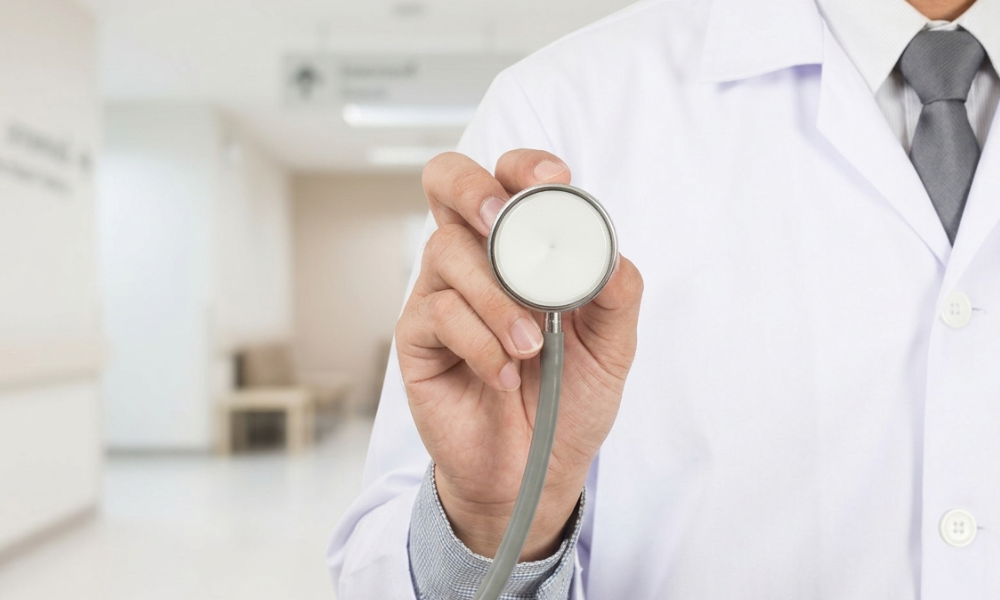 Doctor in a white coat holds a stethoscope toward the camera in a bright clinic hallway, signaling medical care.