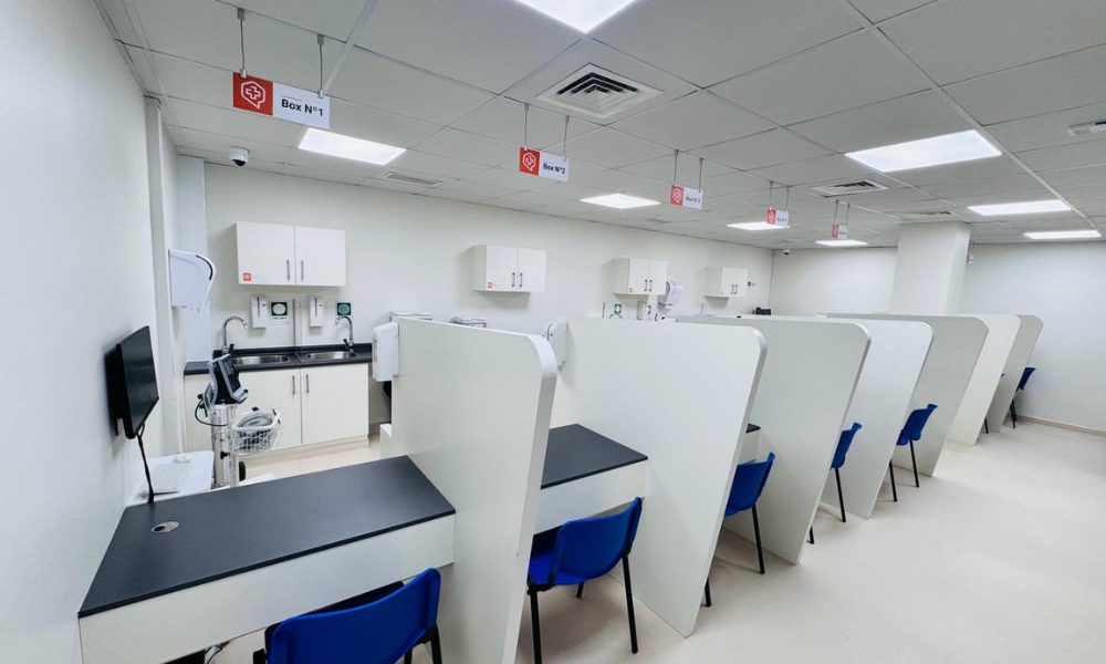 Row of private medical cubicles with blue chairs, service counters, and overhead signs in a clinical setting.