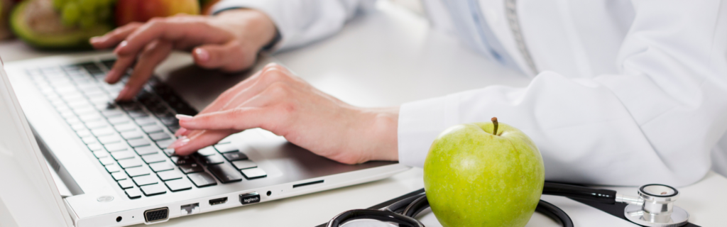 Healthcare professional's hands typing on a laptop; stethoscope and a green apple sit on the desk nearby.