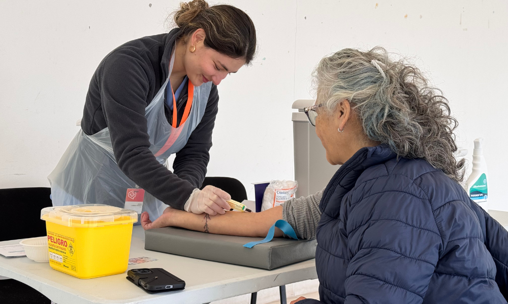 Healthcare worker in a plastic apron and gloves administers a procedure on an older woman’s forearm at a clinic, with a yellow biohazard container nearby.