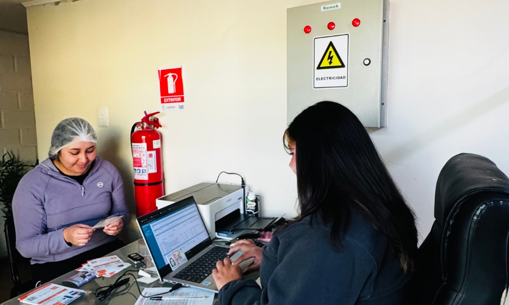 Two women at a desk: one wearing a hairnet and purple jacket reviewing cards while a laptop and printer sit nearby.