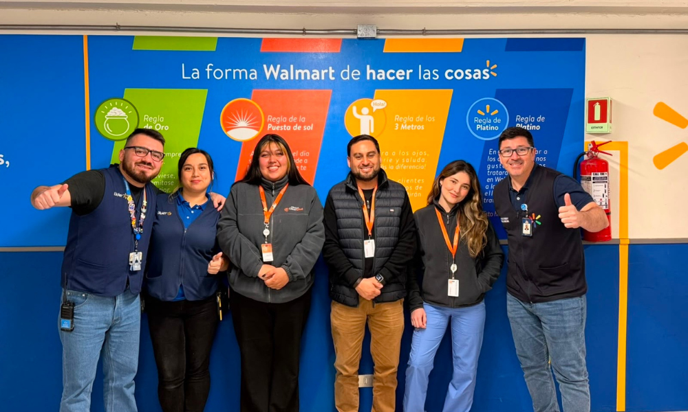 Group of six Walmart employees posing for a photo in front of a colorful wall with the slogan “La forma Walmart de hacer las cosas,” all wearing lanyards and badges.