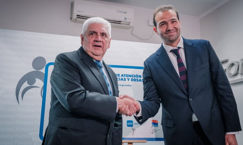 Two men in business suits shake hands in front of a banner with logos, signaling a formal agreement or partnership.
