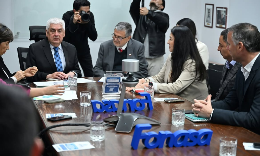 Group of professionals in a meeting around a wooden conference table; blue 'Fonasa' sign sits in foreground, photographers in background capturing the moment.