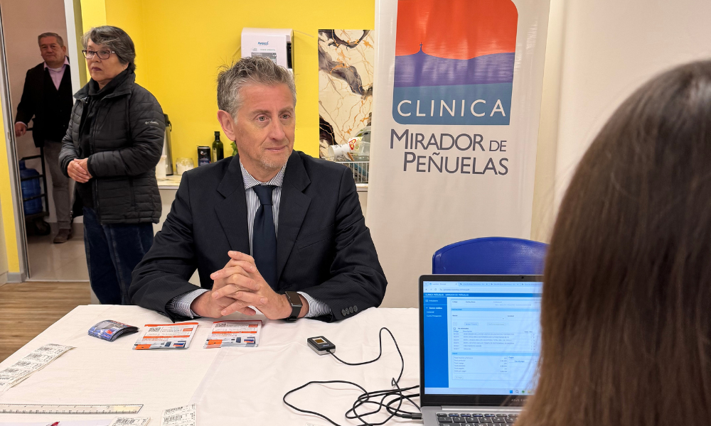 Man in a dark suit sits at a table with pamphlets at a clinic information booth; a banner behind reads ‘CLINICA MIRADOR DE PEÑUELAS’ with a laptop on the table in the foreground.