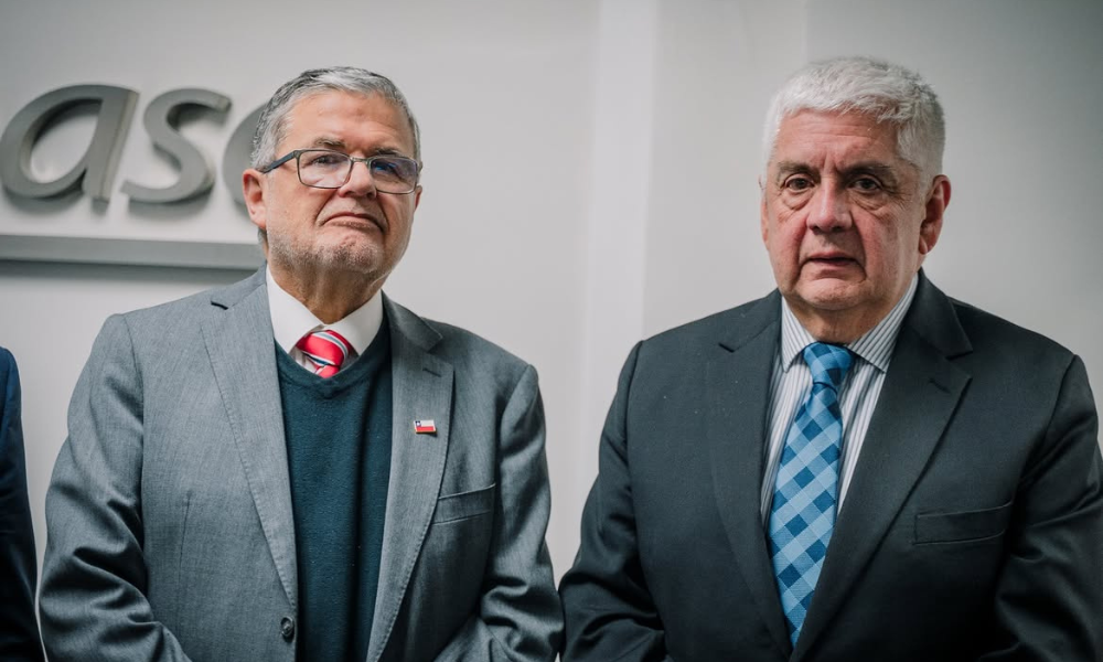 Two older men in suits standing side by side at a formal event, looking toward the camera in a neutral pose.