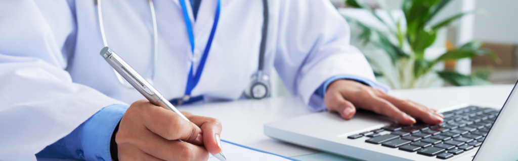 Healthcare professional in a white coat writes with a pen while typing on a laptop, stethoscope around neck