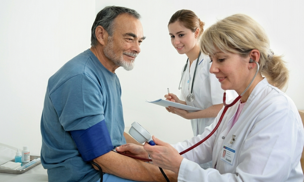 Doctor checks patient’s blood pressure with a cuff while a nurse notes on a clipboard in the background.