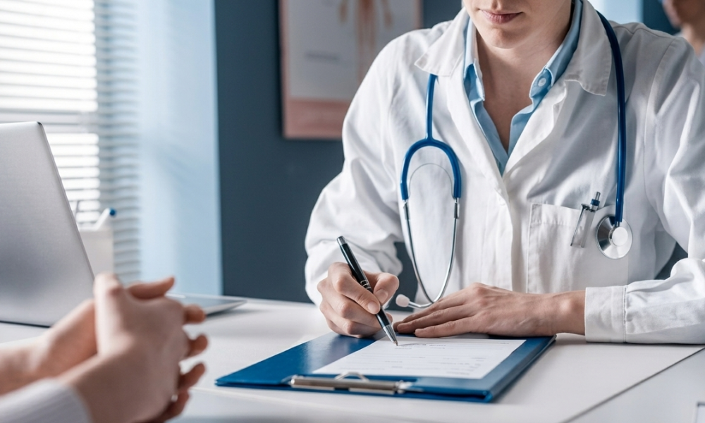 Medical professional in a white coat signs a form on a blue clipboard at a desk, stethoscope around the neck.