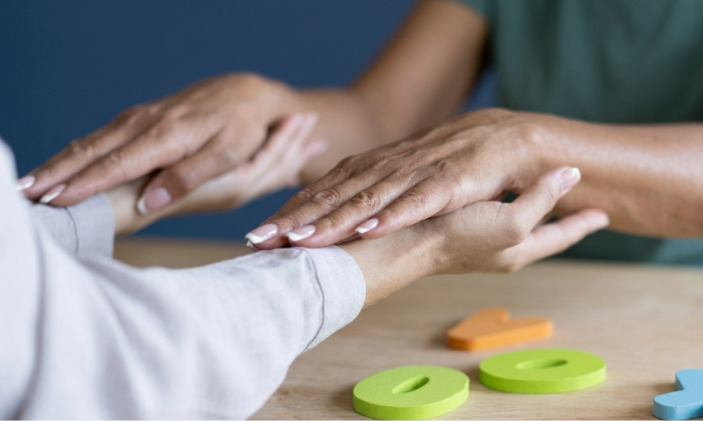 Two people gently hold hands in a touch-therapy or reassurance activity on a wooden table with colorful wooden pieces nearby